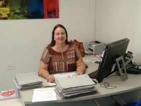 Imagem da diretora do PROCON de Campinas, Dra Yara Pupo, sentada em sua mesa. Na mesa processos, seu computador e ao fundo uma impressora. A diretora está sorrindo para a foto.