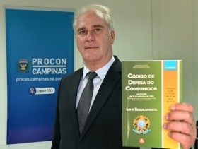 Foto do novo diretor do PROCON Campinas em frente a um banner da instituição e segurando um exemplar do código de defesa do consumidor.